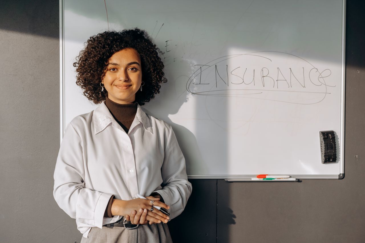 services-04 Smiling woman with curly hair standing by a whiteboard with insurance written on it, conveying professionalism.